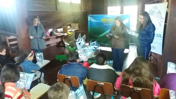 A foto mostra três professoras em pé, lendo a cartilha em quadrinhos "Graxa e sua Turma" para um grupo de alunos, cada qual com um exemplar em mãos, sentados em suas classes. O display com os personagens está ao lado das professoras.