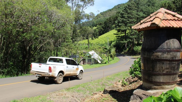 A foto mostra um carro circulando pela estrada conhecida como Caminho das Pipas. A paisagem é repleta de árvores e vegetação. Em primeiro plano, próximo à estrada, uma pipa colonial, alusiva à produção de vinhos em cantinas.