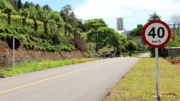 A foto mostra a rodovia Caminho das Pipas, em Rolante, recém asfaltada, com faixa central pintada. Ao lado direito há uma placa com limite de velocidade de 40 quilômetros por hora. No outro lado, um barranco com um parreiral no topo e igreja ao fundo.