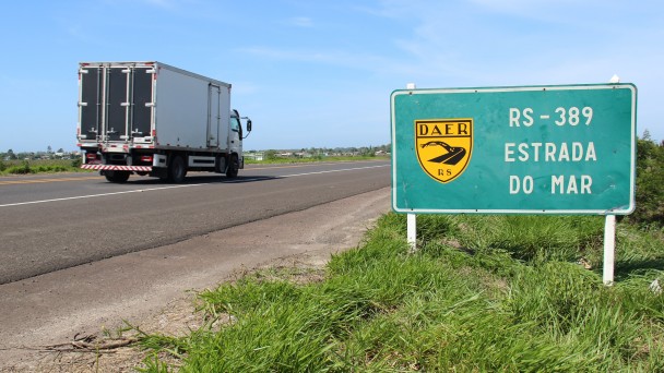 A foto mostra, em primeiro plano, a placa de trânsito que identifica a rodovia ERS-389, conhecida como Estrada do Mar. Ao fundo, aparece a rodovia com um caminhão circulando.