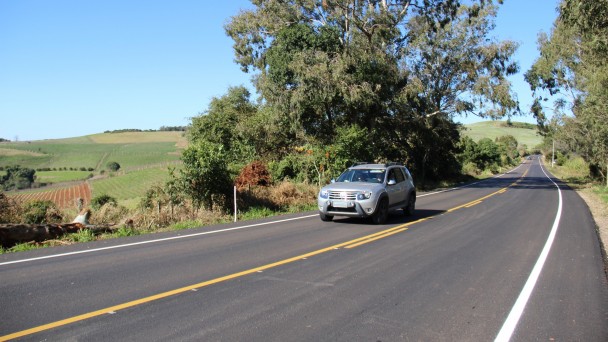 Na foto, um carro circula pela ERS-431, que está com asfalto e pintura da pista em condições renovadas. Ao fundo, árvores e parreirais compõem a paisagem, que tem um céu azul e limpo.