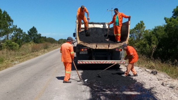 A foto mostra quatro servidores do Daer, uniformizados com macacões laranja, realizando reparos numa rodovia. Eles estão em um caminhão, despejando material asfáltico na estrada.