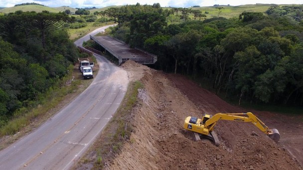 A foto panorâmica mostra a rodovia ERS-332, com a ponte inacabada sobre o Arroio Taipa ao fundo. Mais à frente, na lateral da rodovia, uma retroescavadeira retira terra.