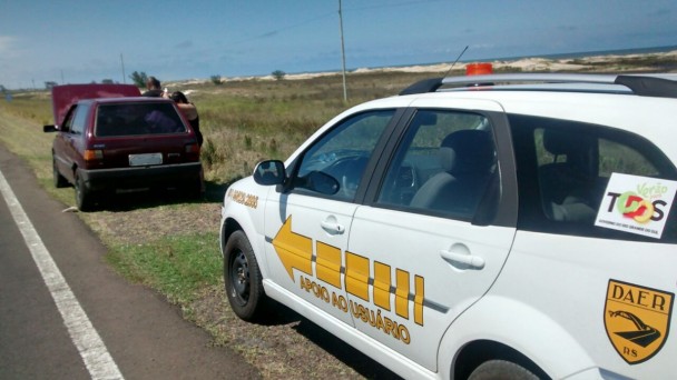 A foto mostra em primeiro plano um carro branco do programa Verão na Faixa, do Daer, estacionado à margem da rodovia. O veículo tem adesivos do Daer, do governo do Estado e marcas do programa em si. Logo à frente, há outro carro, com problema mecânico.