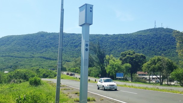 Na foto, um dispositivo OCR, usado no cercamento eletrônico de rodovias, está afixado na beira de uma rodovia. Um carro branco passa pelo equipamento.