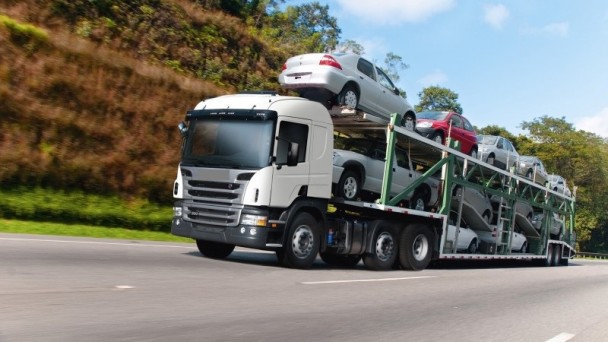 A foto mostra um caminhão cegonheiro que carrega automóveis circulando em uma rodovia.