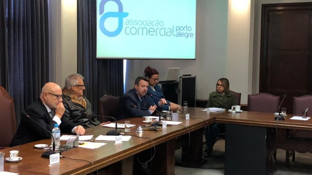 A foto mostra uma mesa quadra onde estão sentados os diretores da Associação Comercial e o diretor do DAER, Lauro Hagemann. O diretor Hagemann está falando sobre a rodoviária. Ao fundo, uma tela com o nome Associação Comercial de Porto Alegre.