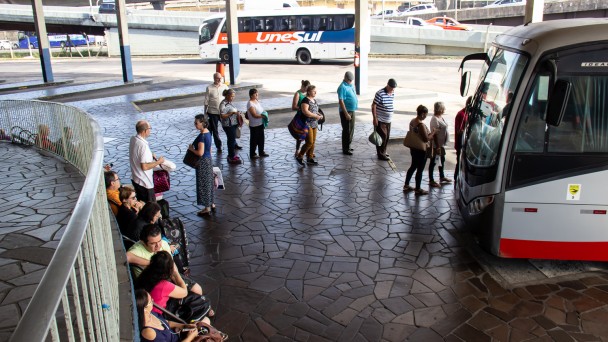 A imagem mostrar um ônibus de transporte intermunicipal na rodoviária de Porto Alegre. Nela, aparecem passageiros na fila para embarcar e outras pessoas sentadas nos bancos da rodoviária.