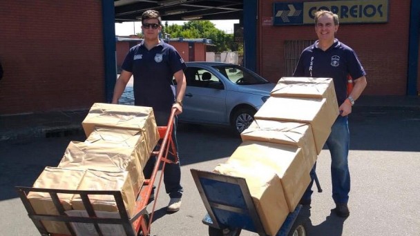 A foto mostra dois servidores transportando os kits de proteção contra o coronavírus. Eles seguram carrinhos com pacotes em frente a rodoviária de Porto Alegre.