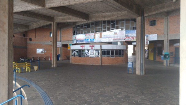 Na foto aparece a faixada de uma rodoviária vazia, com os guichês de atendimento. Ao redor da estrutura estão presentes bancos para se sentar e telefones colocados nas paredes.