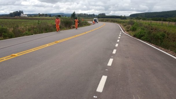 Na foto aparece uma estrada pavimentada em meio a vegetações dos dois lados. Na parte da esquerda podem ser vistos trabalhadores utilizando uma roupa laranja e ao fundo de um deles está um veículo viajando pela rodovia.