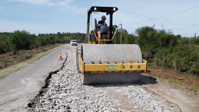 Na imagem aparece está uma máquina de cor amarela passando pela estrada em uma parte que estão dispostas britas. Nos dois lados do veículo estão presentes as vegetações. Ao fundo da imagem pode ser notado um carro viajando pela rodovia.