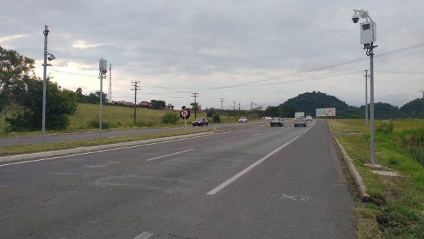 Na imagem pode-se notar a presença de uma estrada, onde estão localizados pardais nos dois lado da foto. Ao fundo do registro estão carros viajando pela rodovia. O céu está nublado, como se estivesse prestes a chover.