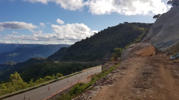 Na imagem pode-se ver montanhas rochosas e a estrada da Rota do Sol mais ao fundo. A imagem é colorida com muito verde da vegetação ali por perto, além do azul do céu, que estava muito bonito no dia. 