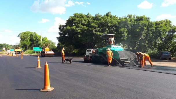 Na foto estão presentes trabalhadores usando laranja, ao redor de uma máquina que está realizando intervenções no asfalto. Cones à esquerda e árvores ao fundo também fazem parte da fotografia.