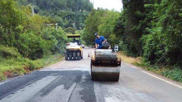 Na foto aparecem duas máquinas para cuidar do asfalto, com os trabalhadores conduzindo. Um deles, o da direita, utiliza uma roupa toda azul. Dos dois lados da imagem, pode-se notar grande presença de vegetação, com várias árvores. 