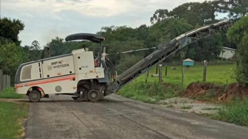 Na imagem está uma grande máquina fazendo intervenções na vegetação próxima da pista. Ela é branca e laranja e tem uma espécie de ganho para alcançar a árvore.