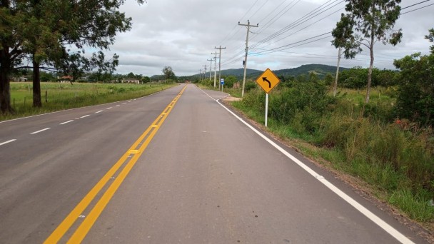 Na imagem aparece a ERS-713 pavimentada com ampla vegetação dos dois lados da rodovia. Na parte direita pode ser vista uma placa amarela de trânsito. 