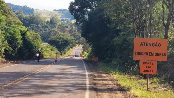 Na foto está presente uma placa laranja na parte direita, onde está escrito "trecho em obras". Nas outras partes da imagem, está a rodovia pavimentada com veículos ao fundo e do lado uma extensa vegetação.