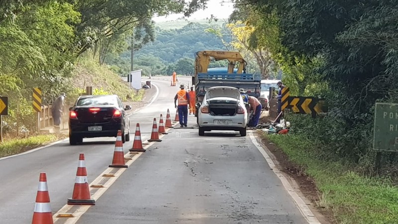 Na foto estão nos centros vários cones laranjas de sinalização. Na parte direita ao fundo, está um carro branco com um caminhão de obras na frente. Dos dois lados extremos da imagem, está uma extensa vegetação.