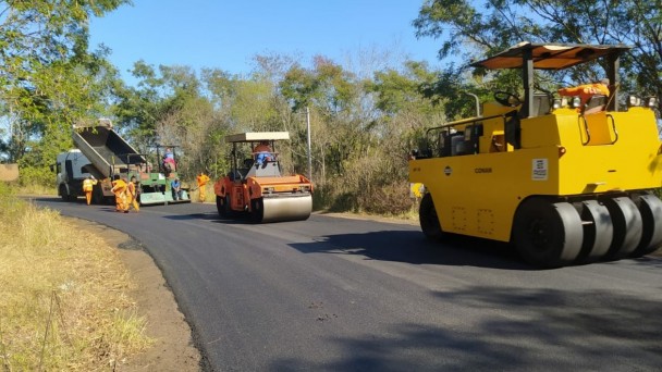 Na foto estão duas máquinas trabalhando no pavimento da estrada. Uma delas é laranja e a outra é amarela. Ao fundo está um caminhão caçamba branco largando material na rodovia.