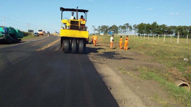 A foto mostra uma máquina na rodovia. Ao redor, funcionários que estão trabalhando na obra.