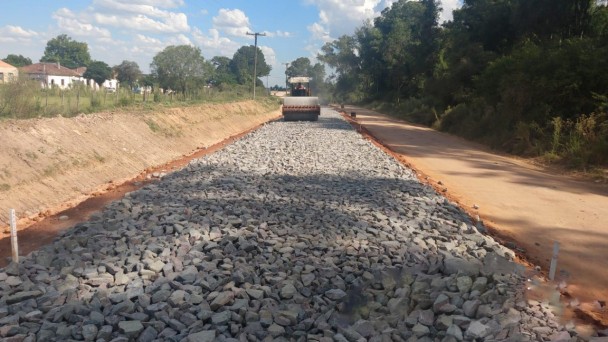 A imagem mostra a máquina realizando a implantação de macadame na estrada.
