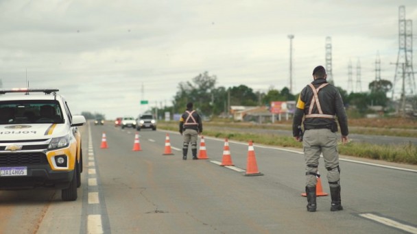 A foto mostra uma viatura do Comando Rodoviário da Brigada Militar estacionada às margens da ERS-118, com dois policiais, junto a cones, fiscalizando o trânsito.