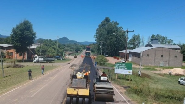 A imagem mostra as máquinas trabalhando no novo asfalto e a placa da obra.