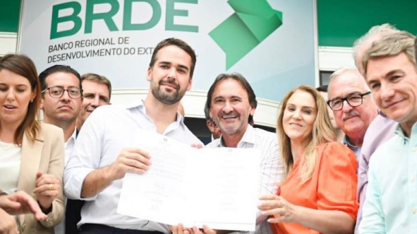 Governador Eduardo Leite e secretário Juvir Costella seguram contrato assinado, com outras pessoas ao lado e marca do BRDE ao fundo.