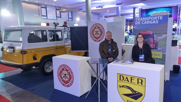 A foto mostra o estande do Daer na Transposul. Nele, há um pupito com logotipo da autarquia e uma mesa de vidro. Dois servidores sorriem para a foto.