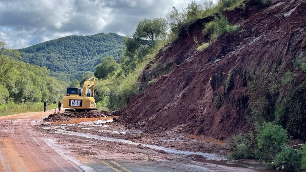 A foto mostra a pista da rodovia com terra e árvores caídas. Nela, um equipamento realiza o trabalho de limpeza.