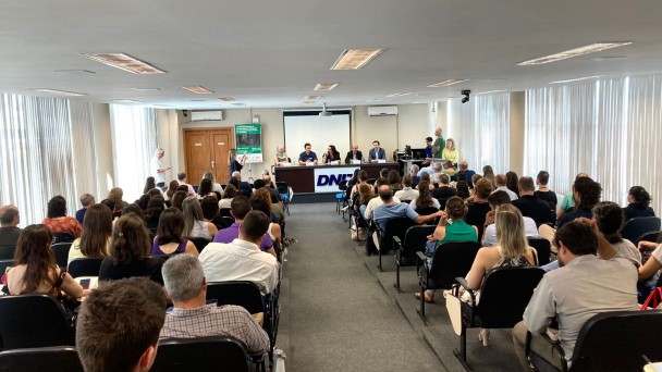 Imagem mostra auditório com pessoas observando palestrantes.