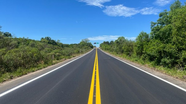 Imagem mostra pista com asfalto recuperado, com árvores nas laterais e céu azul acima.