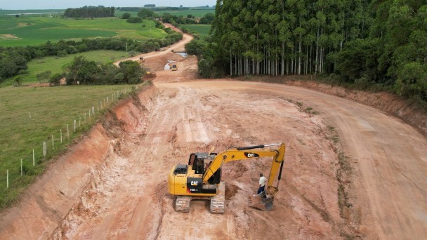 A foto mostra uma retroescavadeira em obras na rodovia de acesso ao município de Amaral Ferrador. A vista é panorâmica, captada por um drone.