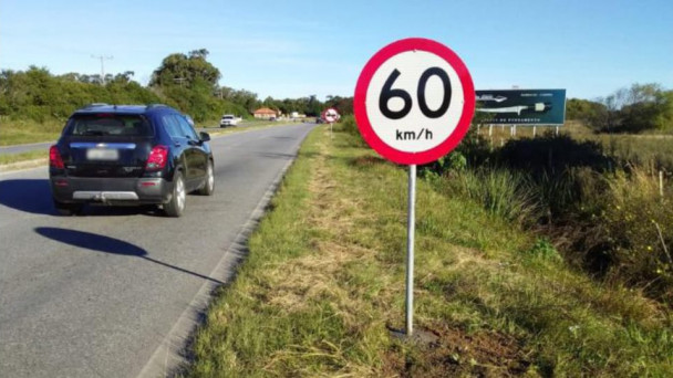 A imagem mostra um carro transitando em uma estrada. Em primeiro plano, uma placa de 60 quilômetros por hora.