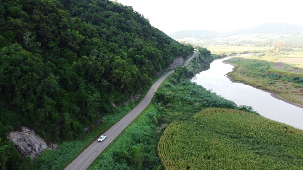 A foto aérea mostra um trecho pavimentado da VRS-811, em Travesseiro. A paisagem tem morros, vegetação densa e um riacho.
