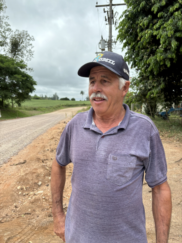 Imagem de um homem com boné preto, camiseta roxa. Está no acostamento da estrada em obras.
