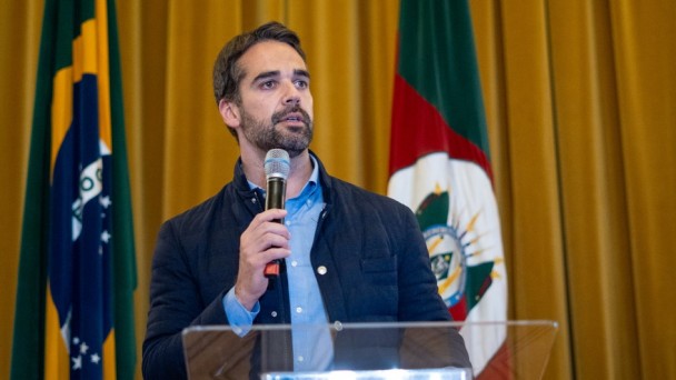 Governador Eduardo Leite discursa, segurando microfone. Está com camisa azul e jaqueta azul marinho.