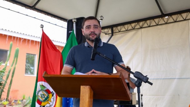 Gabriel Souza, governador em exercício, fala na tribuna ao microfone, usa camiseta azul marinho com listras brancas nas mangas e na gola.